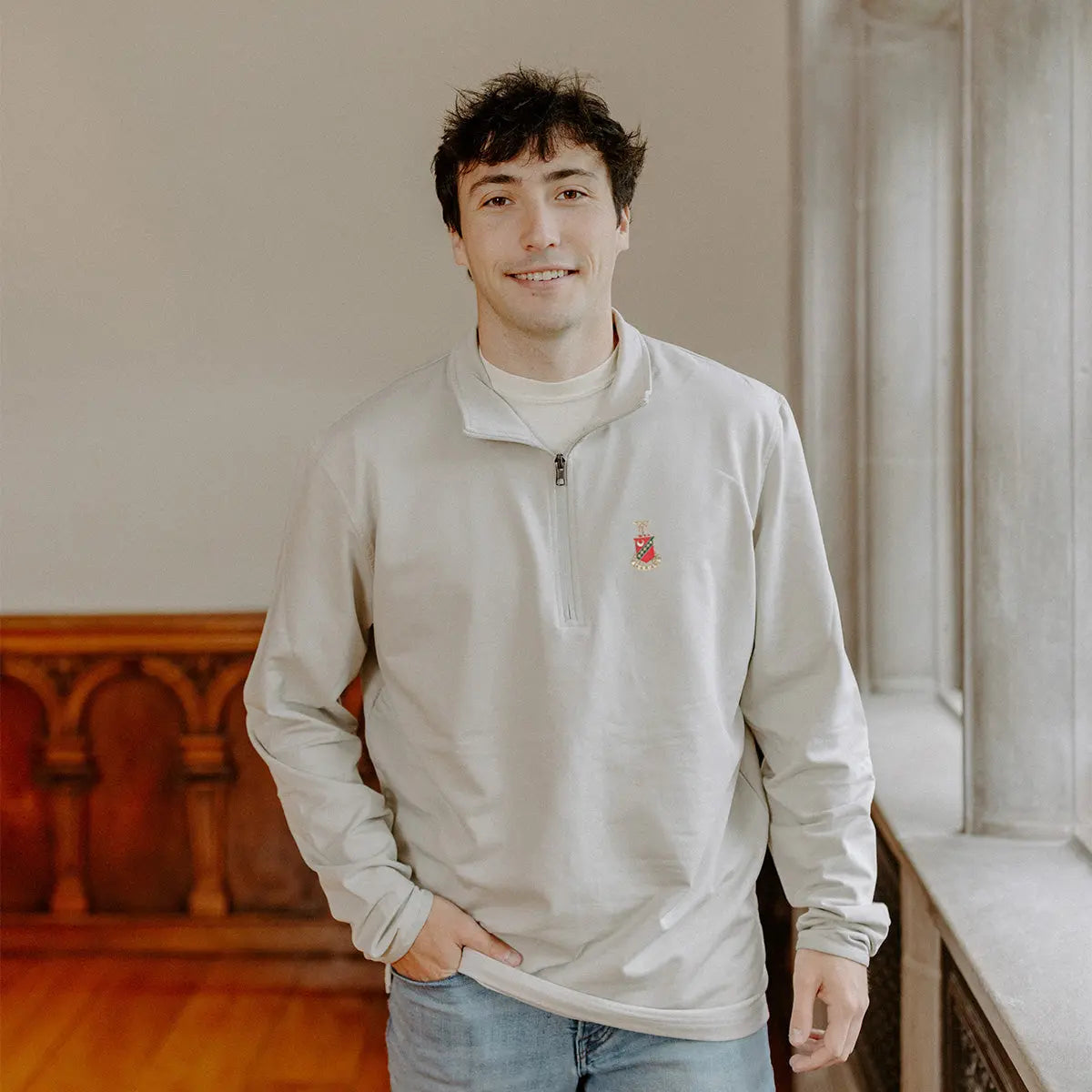 Male model wearing the smoke-colored Kappa Sigma Embroidered Luxe Quarter Zip with crest embroidery, standing by a window in a formal setting.