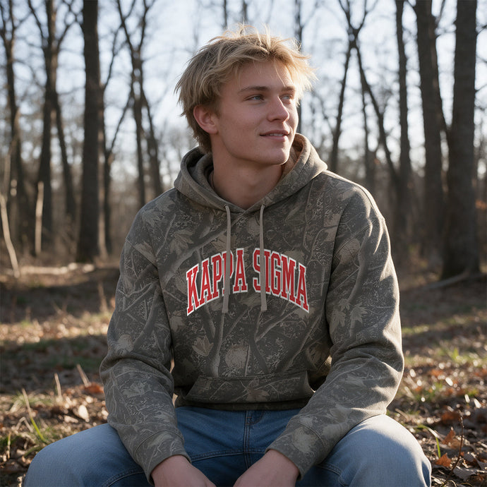 College-aged male wearing the fraternity Deadwood Camo Hoodie with red and white Kappa Sigma lettering, seated outdoors in a wooded setting.