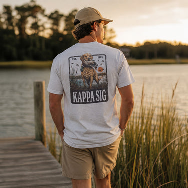 Young man wearing a white graphic T-shirt while standing on a wooden dock overlooking a lake at sunset.