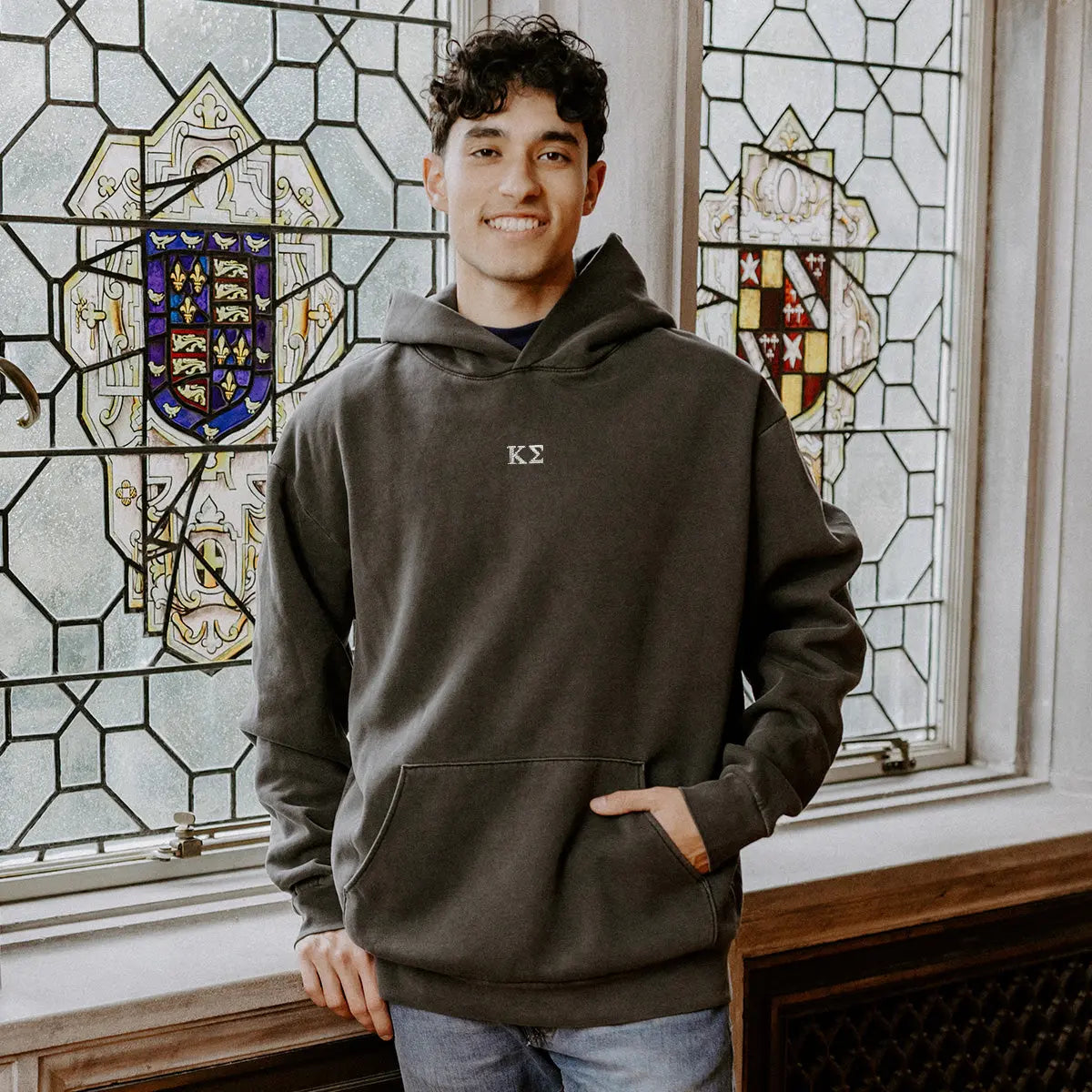 College-aged man wearing the Kappa Sigma embroidered black hoodie, standing in front of stained glass windows with fraternity-style crest designs.