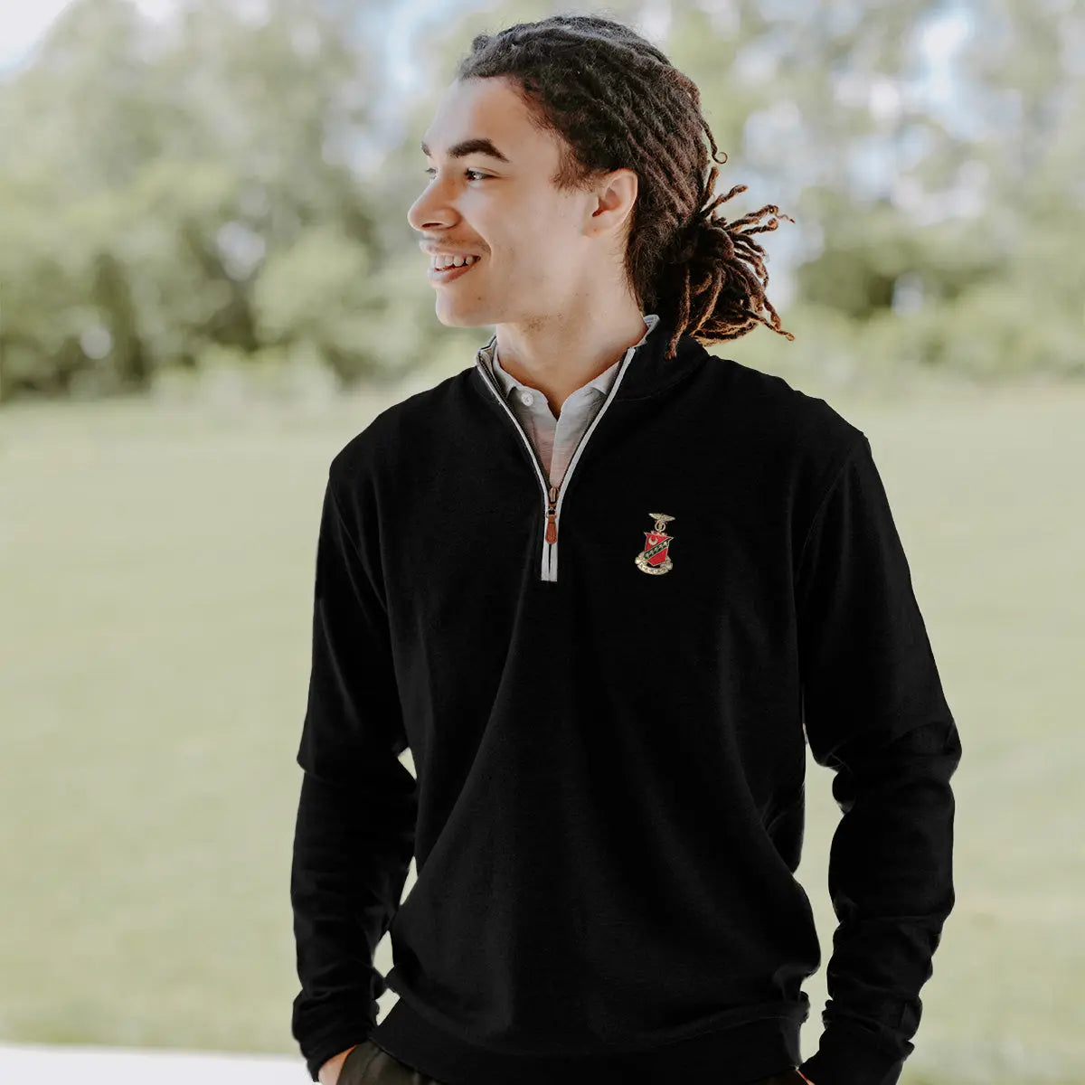 College-aged man smiles and looks off to the side while wearing a black quarter-zip pullover embroidered with a fraternity crest. He stands outdoors in a bright, grassy setting.