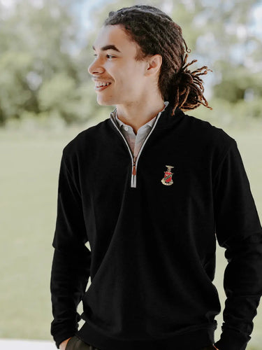 College-aged man smiles and looks off to the side while wearing a black quarter-zip pullover embroidered with a fraternity crest. He stands outdoors in a bright, grassy setting.