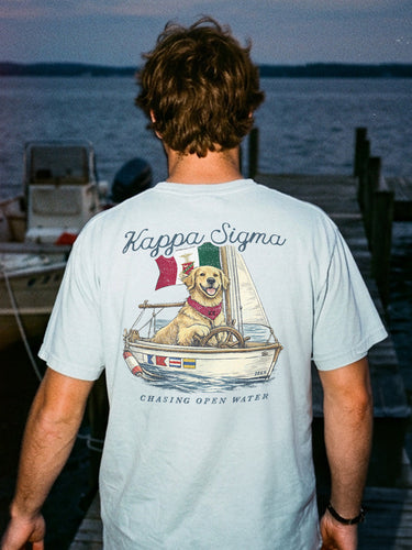 Young man standing on a dock at sunset wearing a light blue t-shirt featuring a golden retriever steering a sailboat with a flag and the phrase “Chasing Open Water” on the back.
