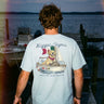 Young man standing on a dock at sunset wearing a light blue t-shirt featuring a golden retriever steering a sailboat with a flag and the phrase “Chasing Open Water” on the back.