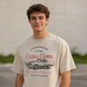 Young man wearing a vintage-style beige graphic T-shirt featuring a retro muscle car illustration and cursive text, standing outdoors with a neutral blurred background.